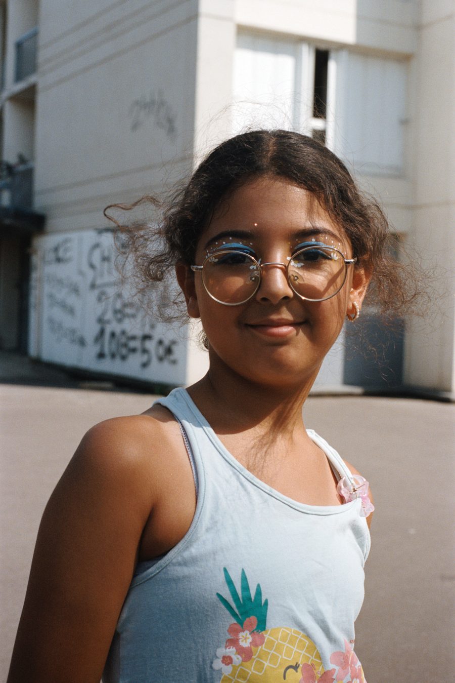 une fille à lunettes, débardeur à ananas, devant les immeubles